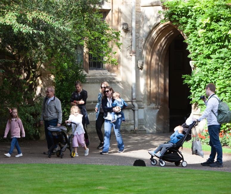 A group of people of various ages walk in a leafy setting with a stone building in the background.