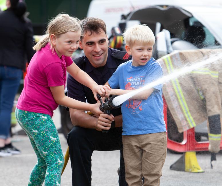 A firefighter crouching to help a boy and a girl direct a firehose.