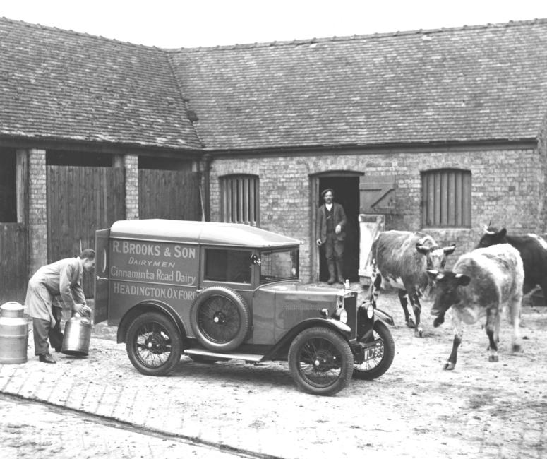 Black and white photograph of a dairyman lifting milk into the back of a van in a farm courtyard. A man standing in a doorway and cows walk by the car.