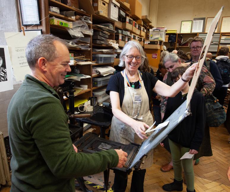 Two people taking a print out of a letterpress with prints pegged onto a line behind.